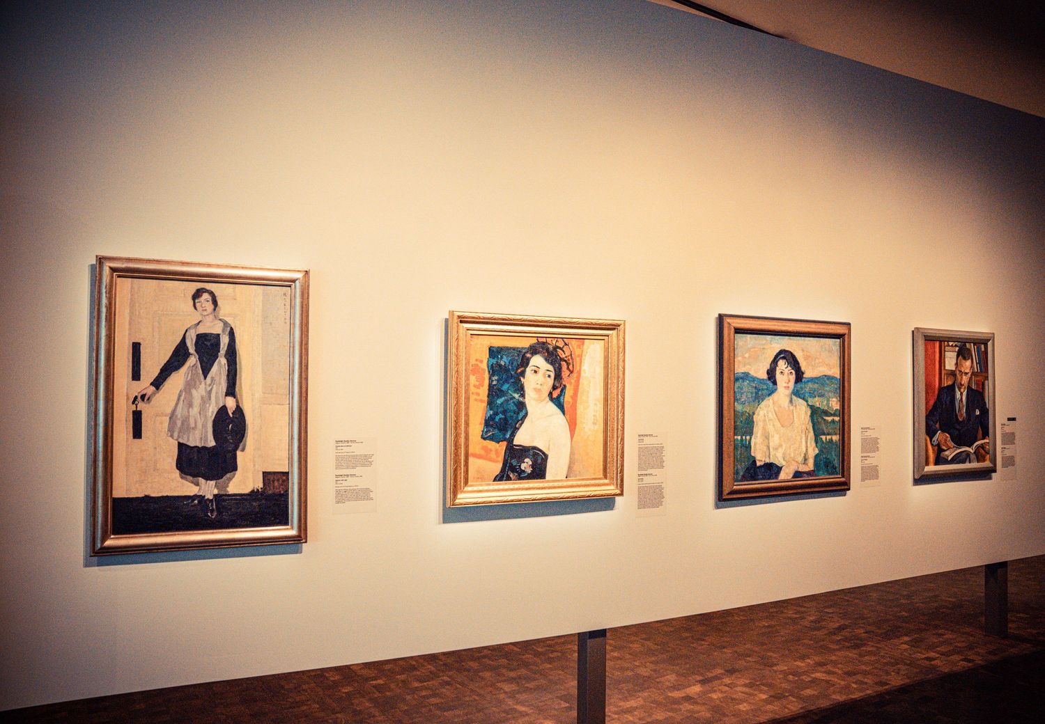 Museum gallery wall with framed portraits arranged in linear sequence under warm lighting.