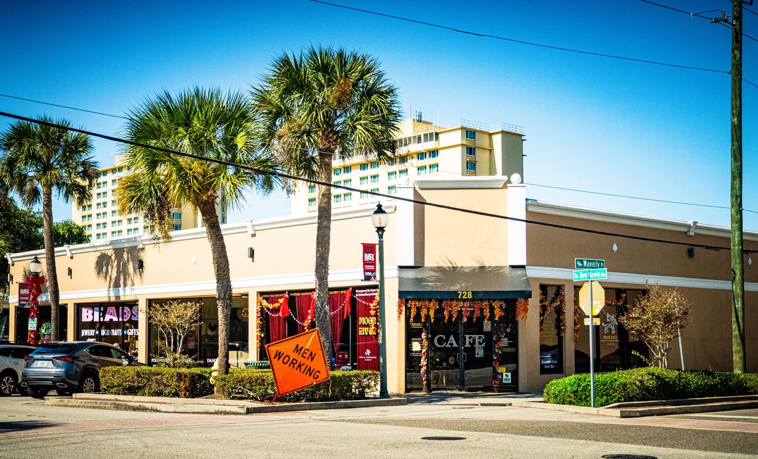 A panoramic view of a strip mall with palm trees and storefronts against a bright blue sky.