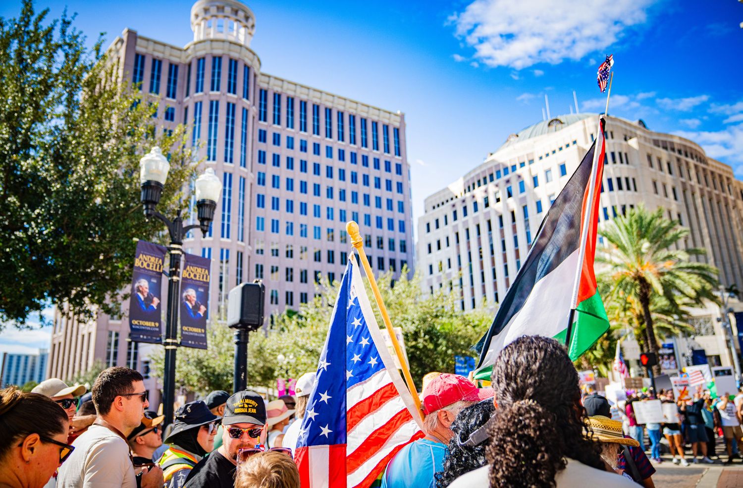 Crowd waving flags and banners during a protest in front of urban buildings.