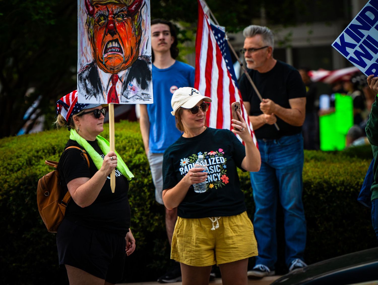 Demonstrators gather on a street with protest signs and American flags during a political rally.