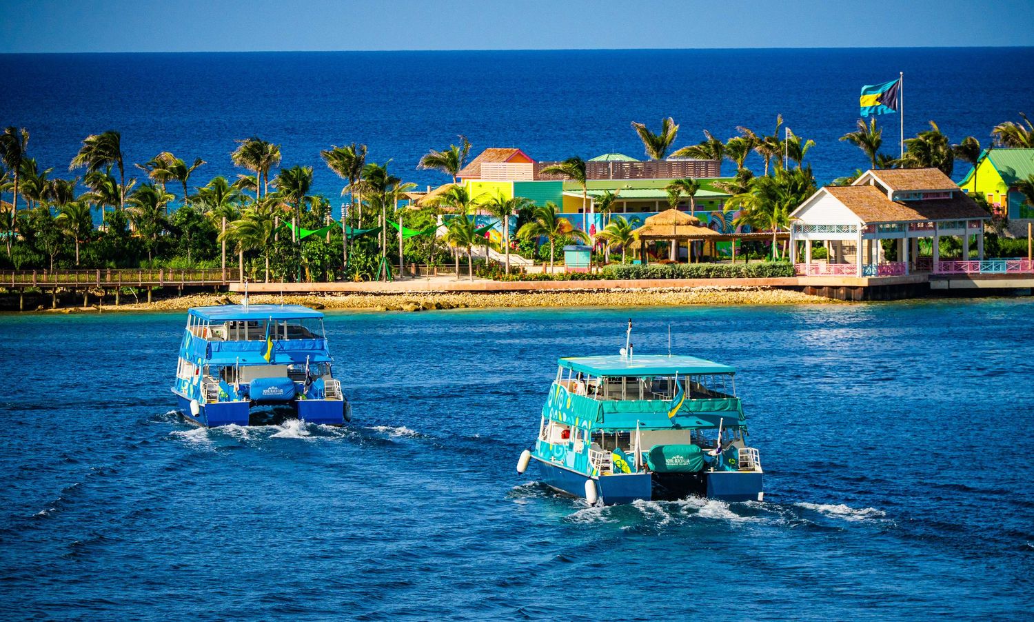 Two colorful tour boats cruise blue waters near a tropical island resort with palm trees and bright buildings.