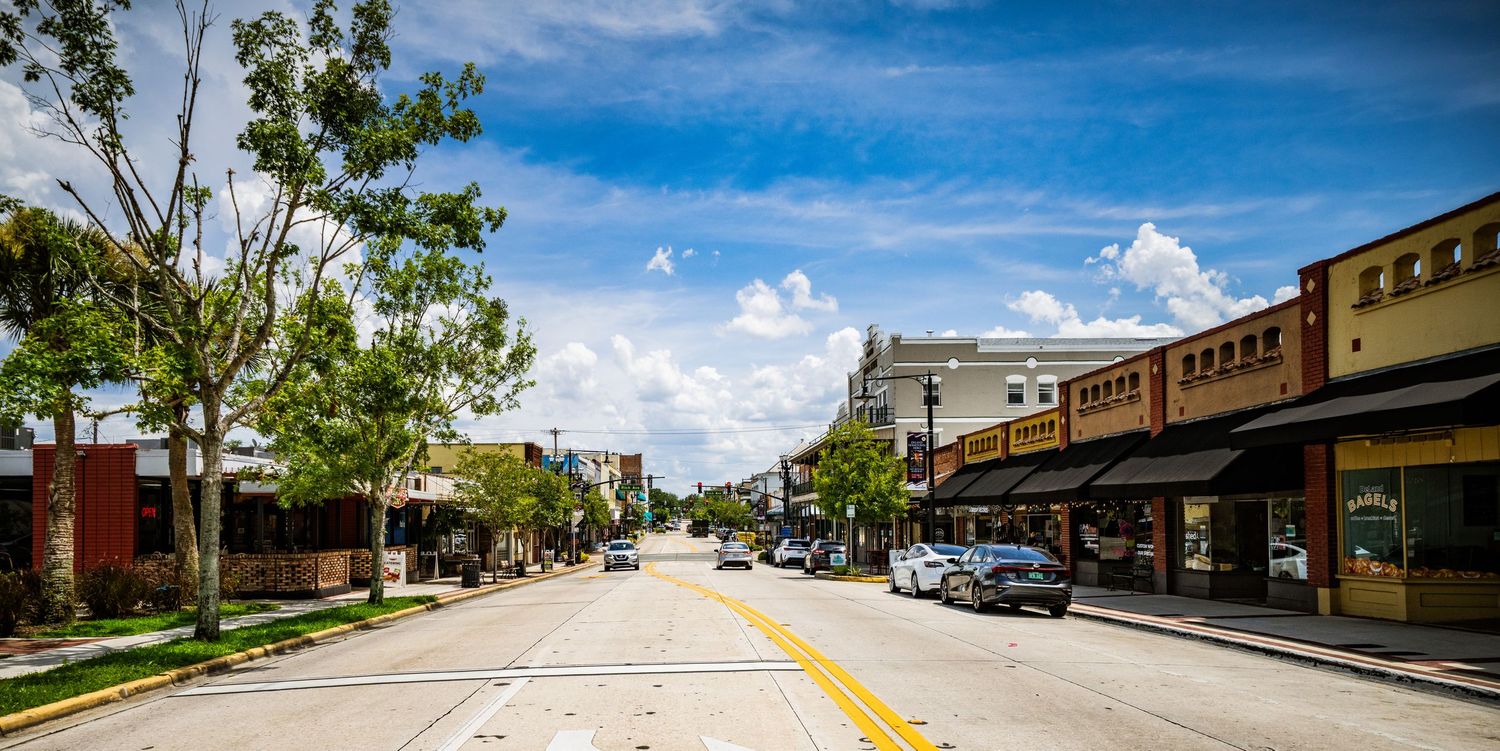 Historic downtown street with brick buildings, parked cars, and trees under blue skies.