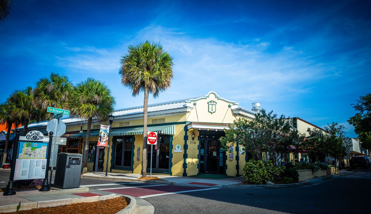 Quaint shopping district with palm trees and Spanish-style architecture under bright blue sky.