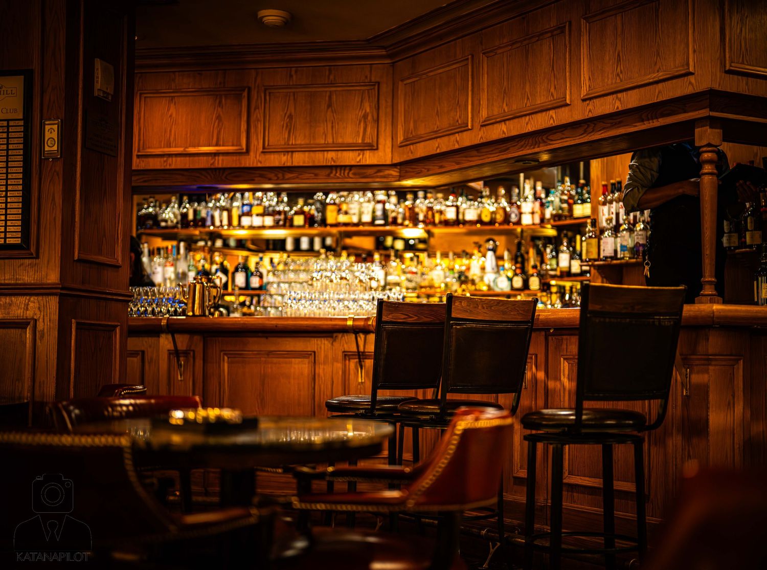 A dimly lit classic wooden bar interior with leather chairs and shelves of illuminated liquor bottles behind the counter.