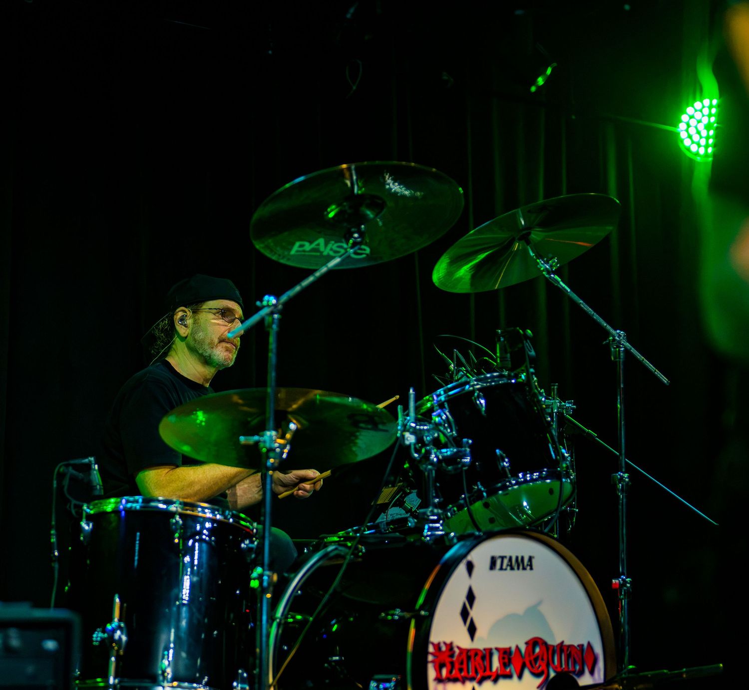 Drummer performing on a Tama drum kit under green stage lighting at a live music concert.