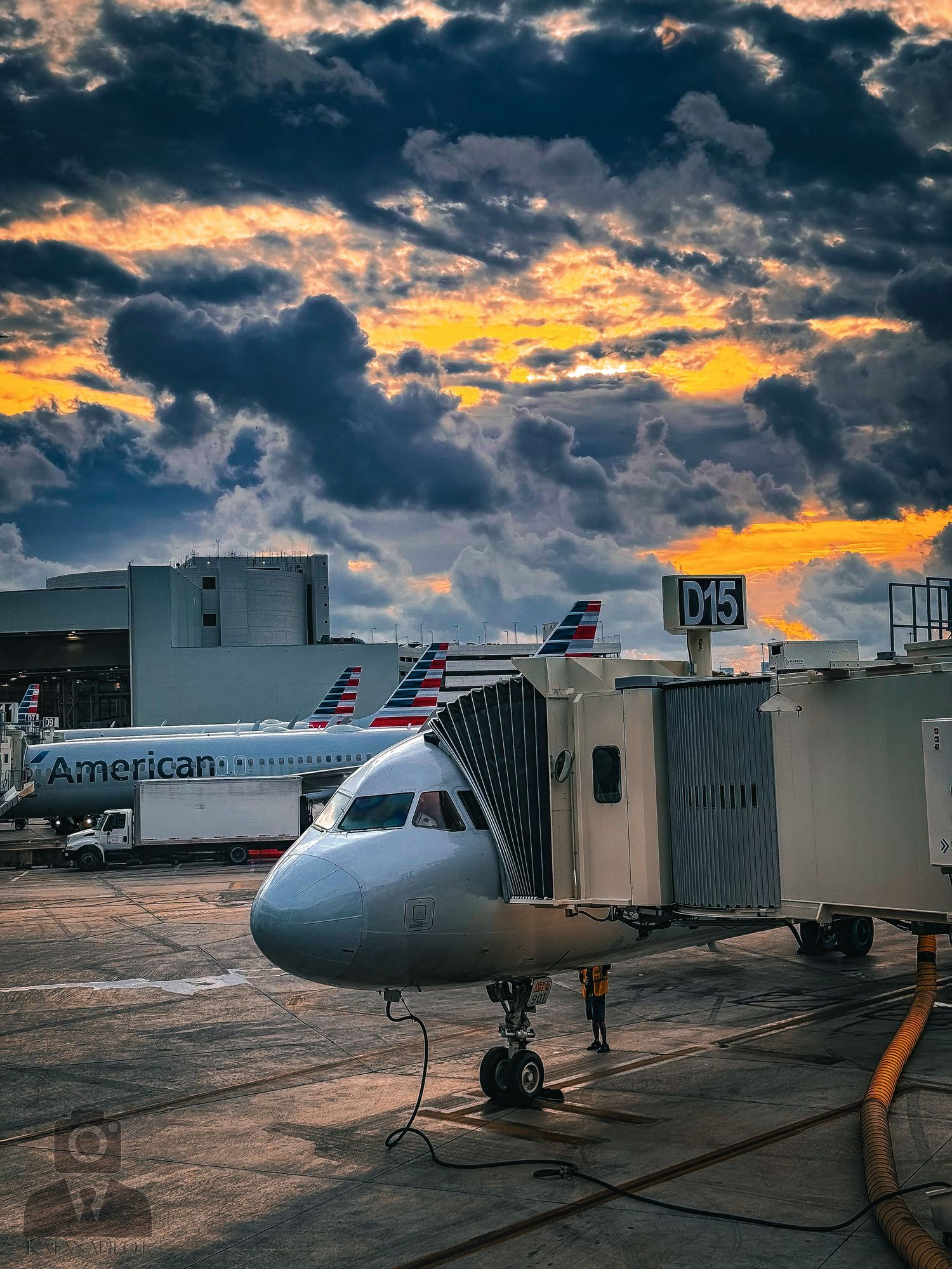 American Airlines aircraft parked at terminal gates during dramatic sunset with orange clouds.