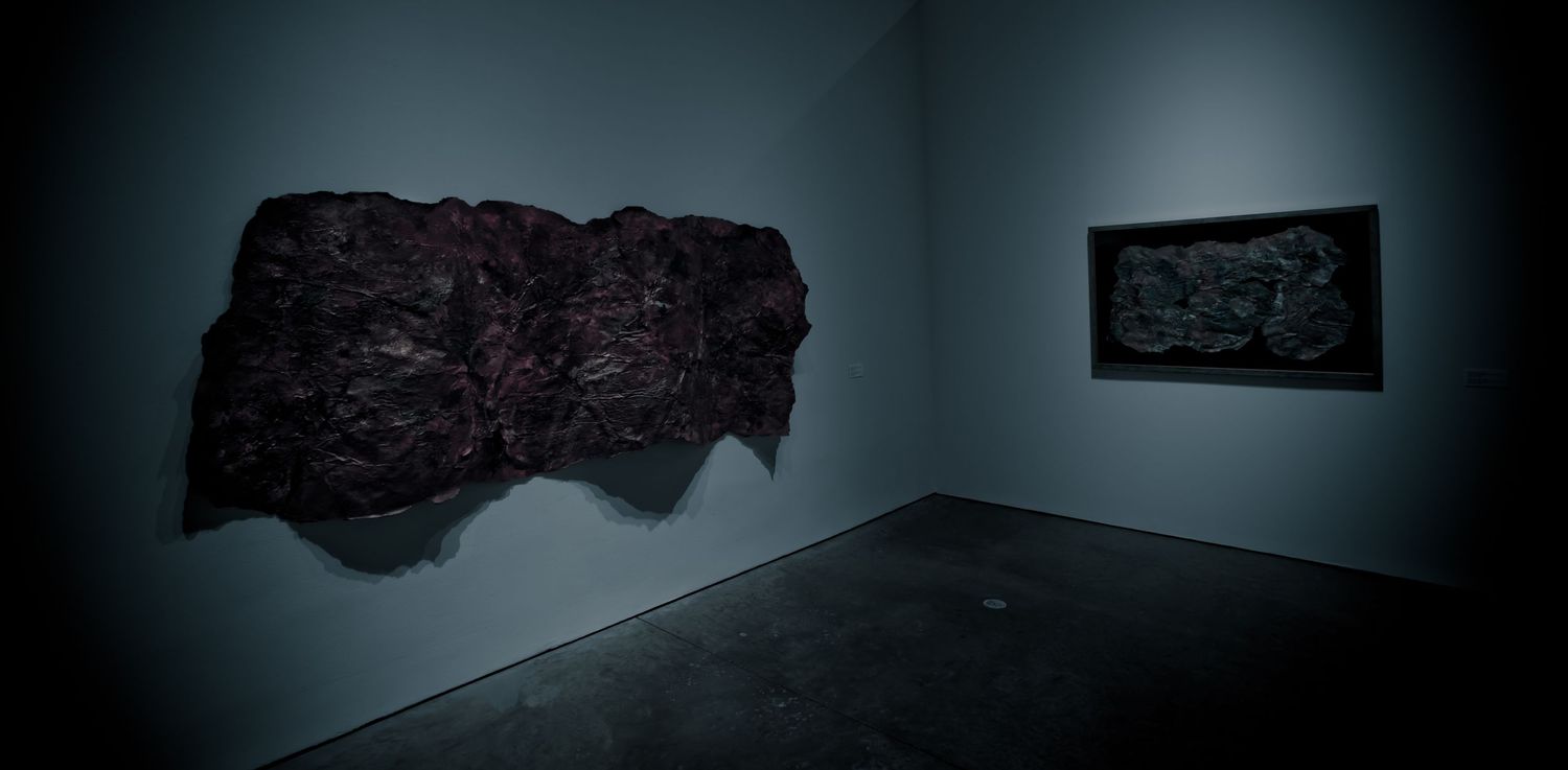 Dark gallery space featuring a textured black wall sculpture and a framed dark artwork on a gray wall.