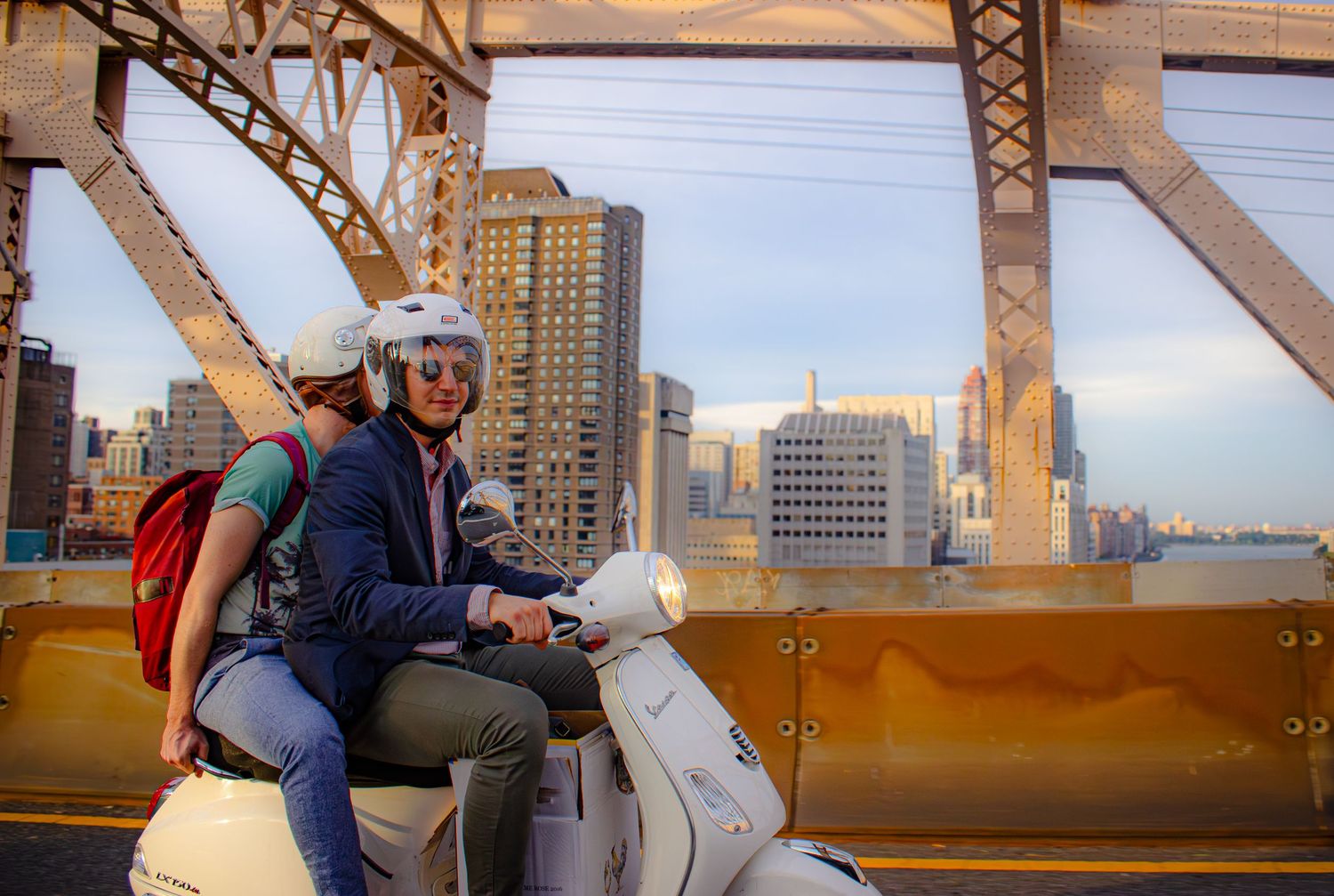 A scooter rider crosses an urban bridge with tall buildings and city architecture visible in the background.