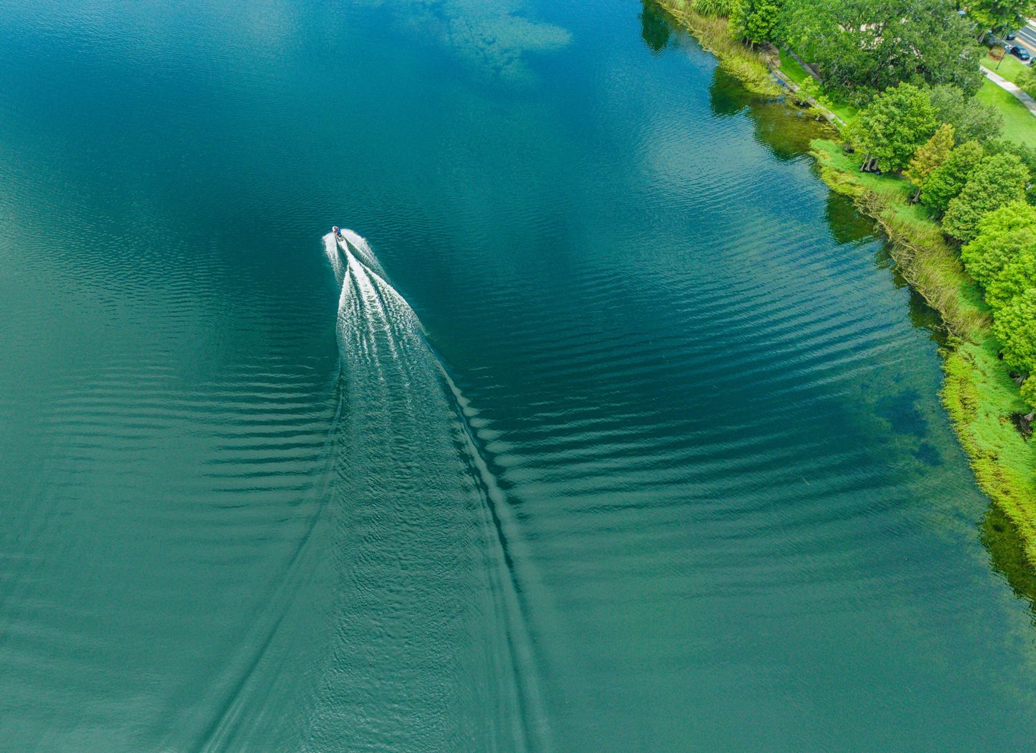 Aerial view of a motorboat creating a V-shaped wake on a serene turquoise lake bordered by lush green trees.