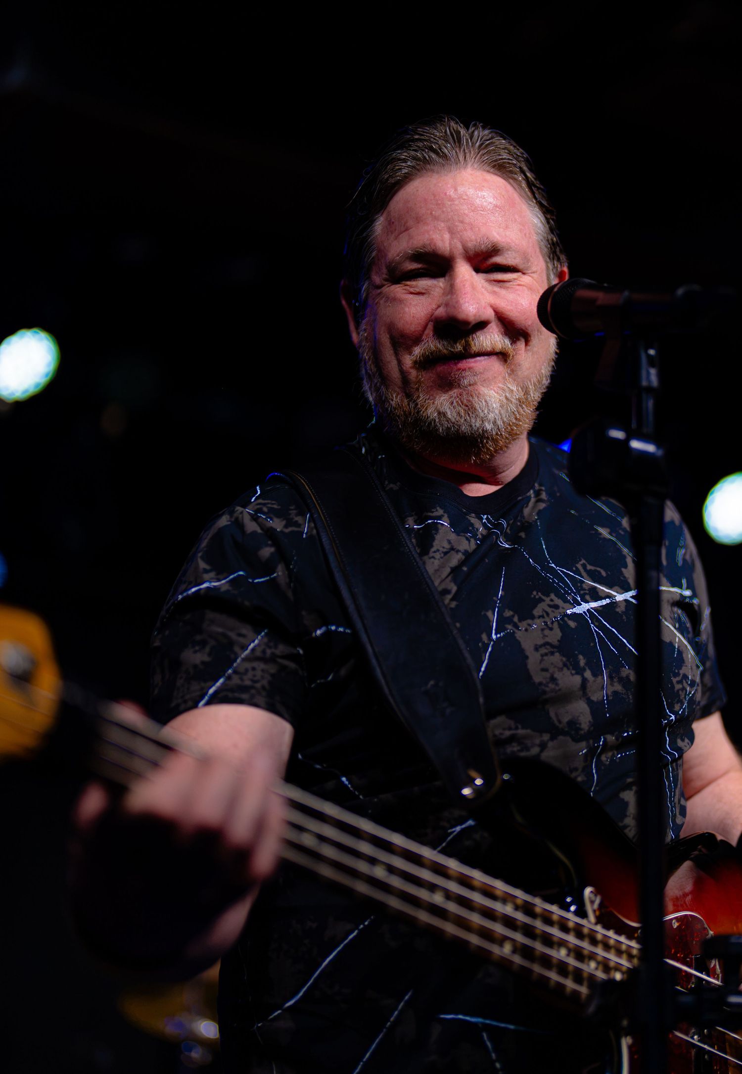 Smiling male guitarist performing live on stage wearing a dark patterned shirt while playing bass guitar.