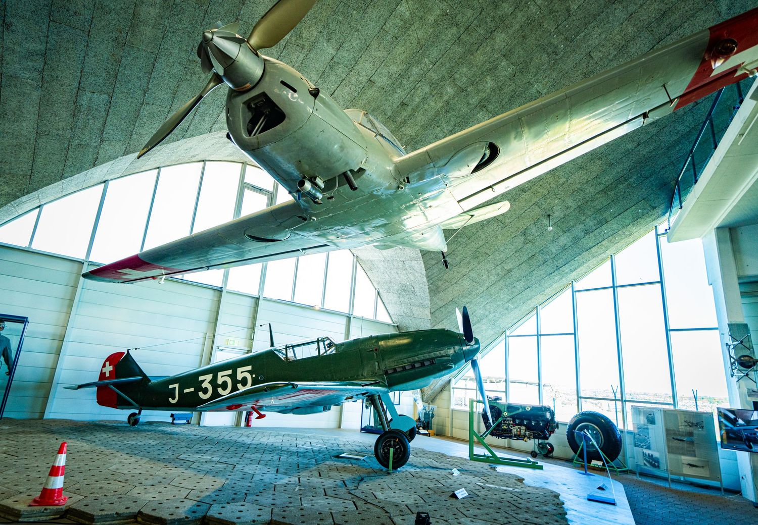 Vintage military aircraft on display in a bright museum hall, with a monoplane suspended overhead and a red J-355 biplane below.
