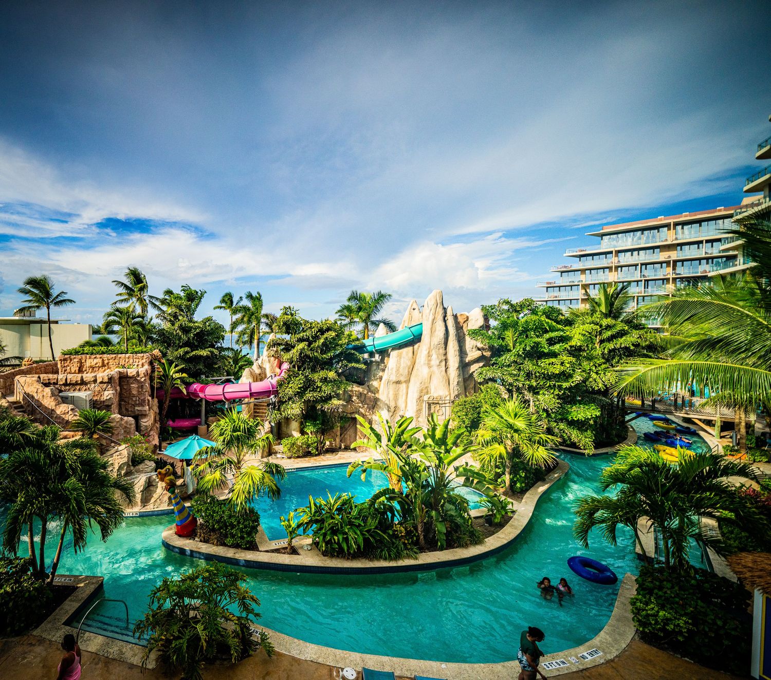 Tropical resort water park with colorful slides, lazy river, lush palm trees, and a hotel backdrop under a vibrant blue sky.