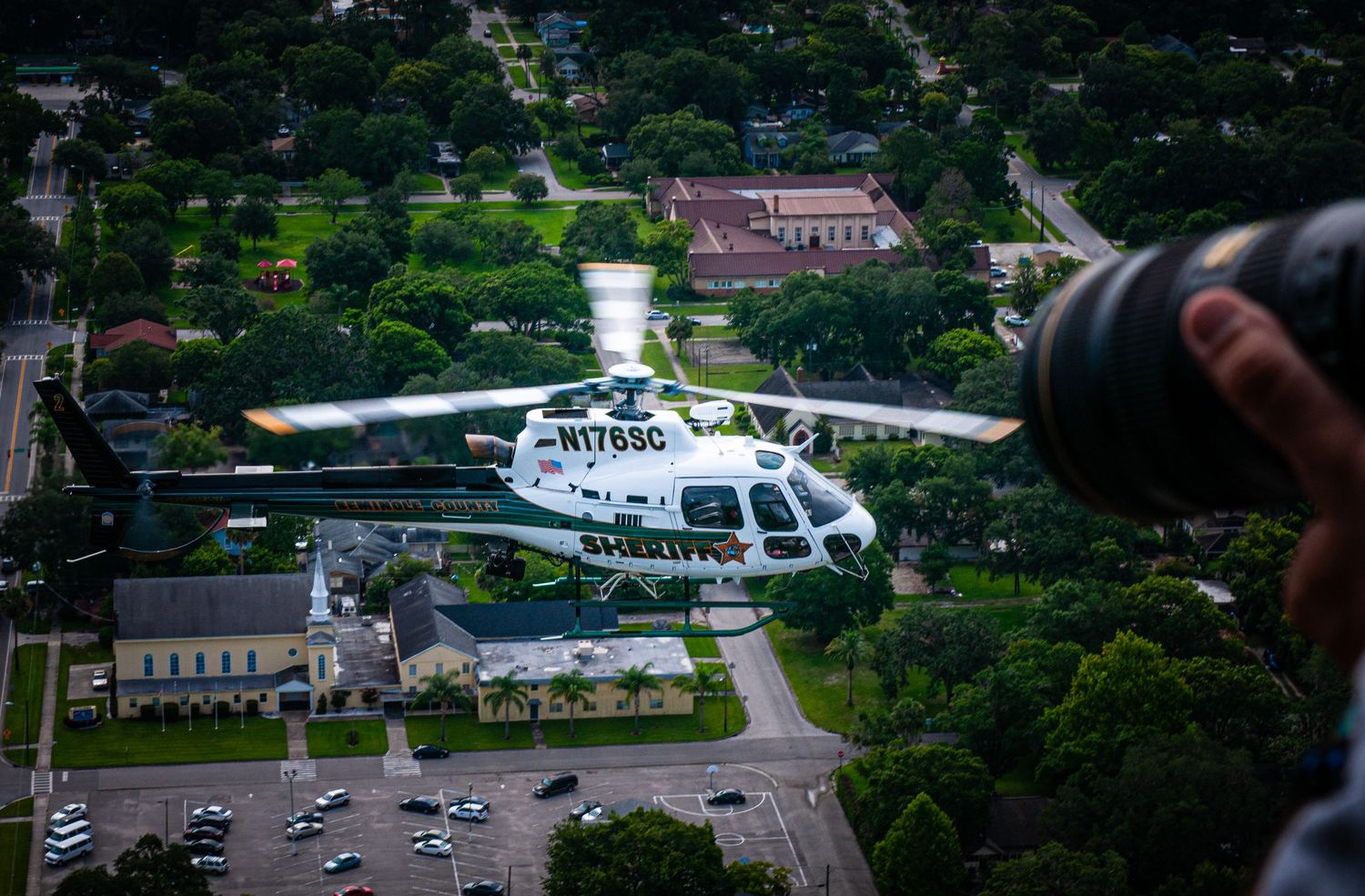 White helicopter flying over suburban area with buildings and trees visible below.