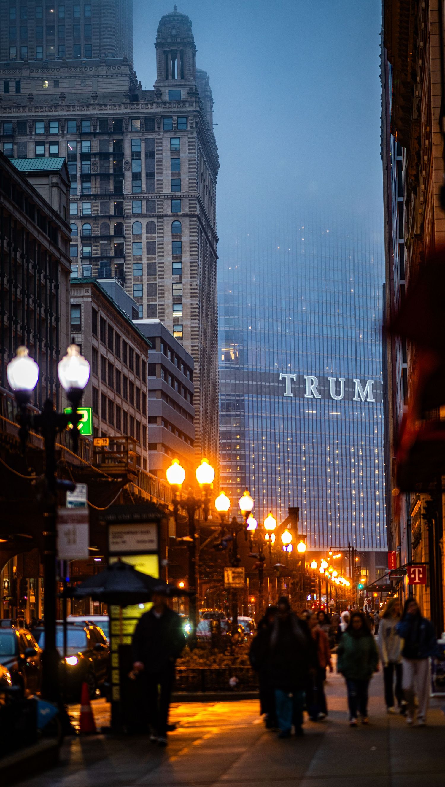 City street at dusk with illuminated lamp posts and historic buildings against a dark urban skyline.