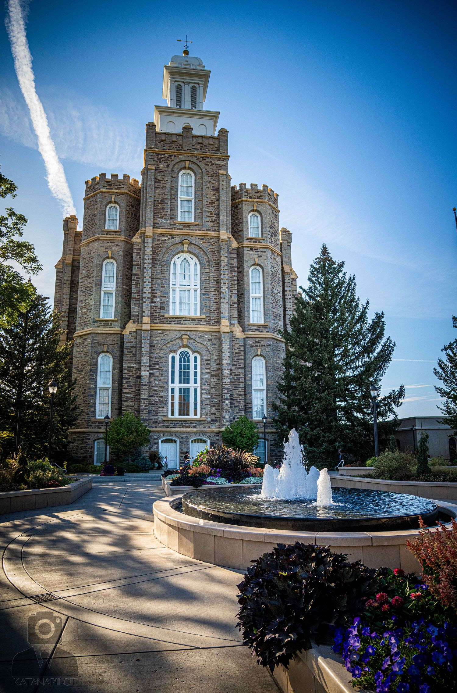 Majestic stone temple with circular fountain feature and colorful landscaping in foreground.