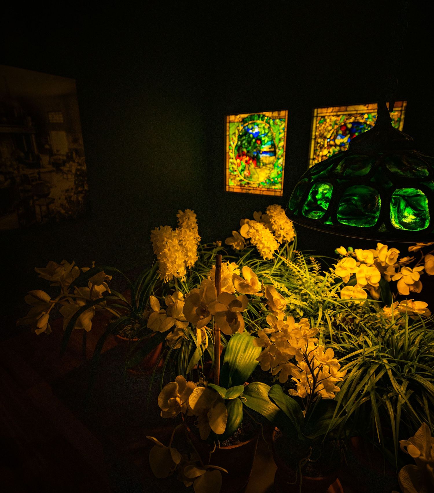 Display of floral arrangements beneath illuminated stained glass windows in a darkened gallery space.