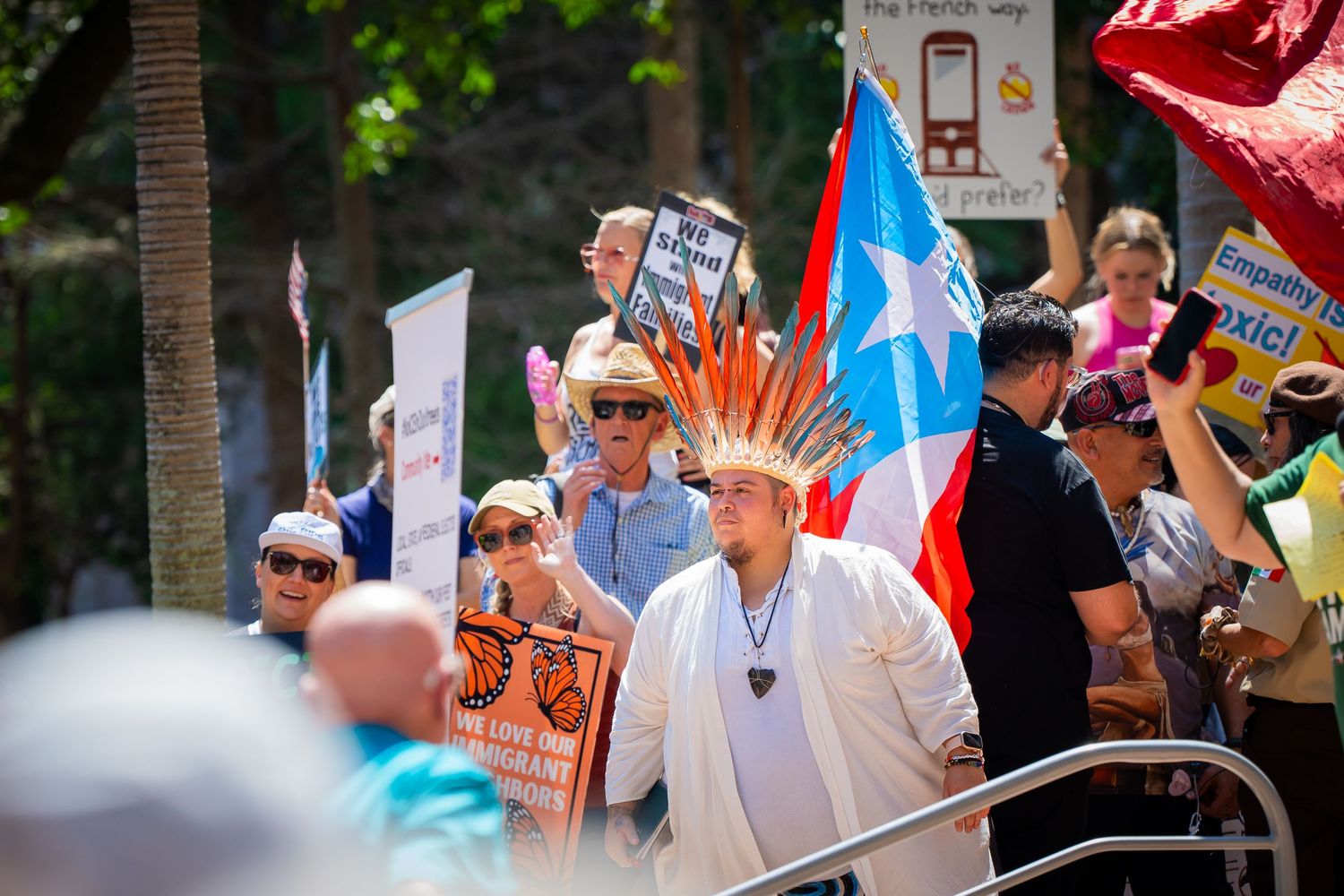 A crowd gathers at a festive outdoor event with colorful banners and flags waving in the sunlight.