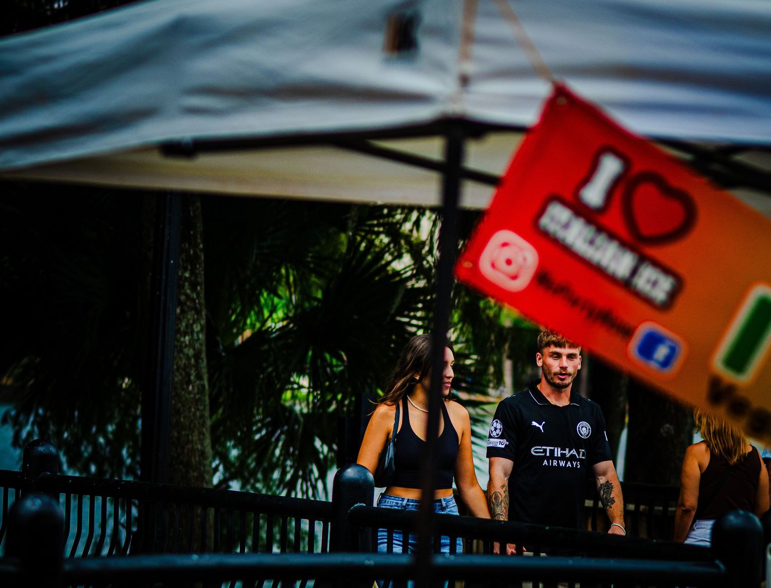 Two people stand under a white tent near a red flag reading I Love at an outdoor event with lush greenery.