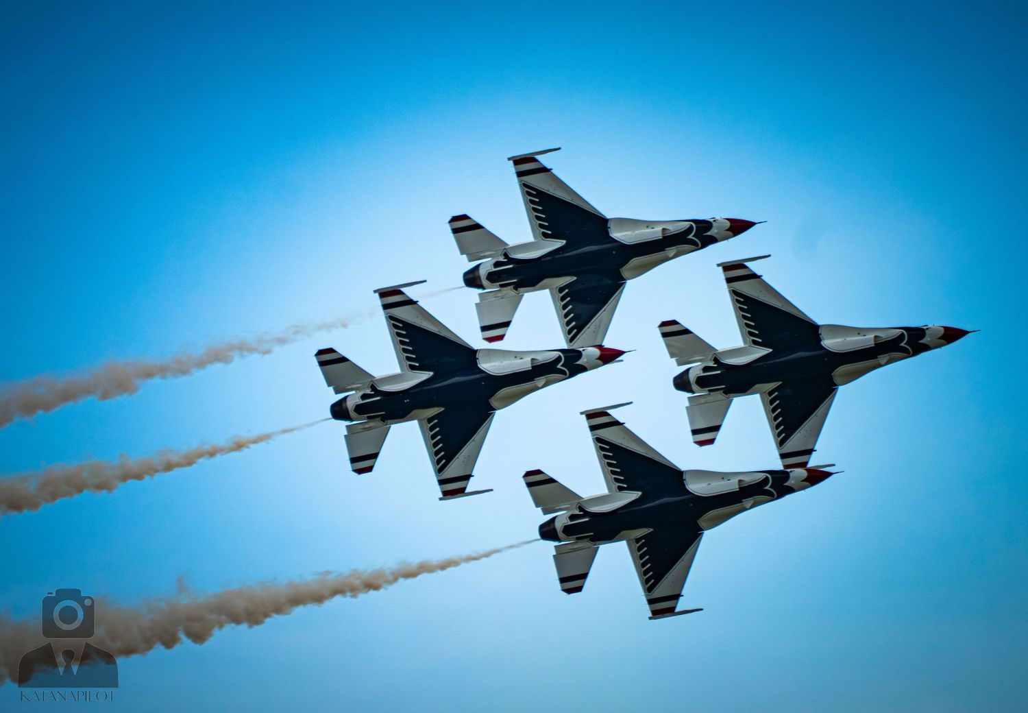 Four military jets fly in tight diamond formation, trailing white smoke against a clear blue sky at an airshow.