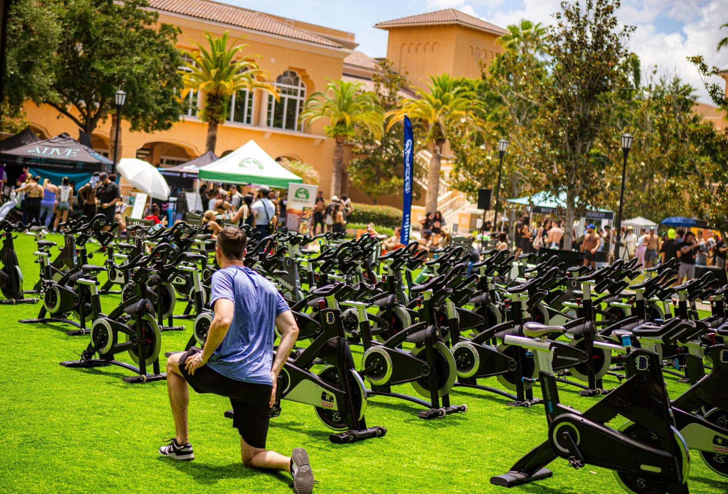 Outdoor fitness event with rows of stationary bikes on green turf, man stretching in foreground, colorful tents in background.