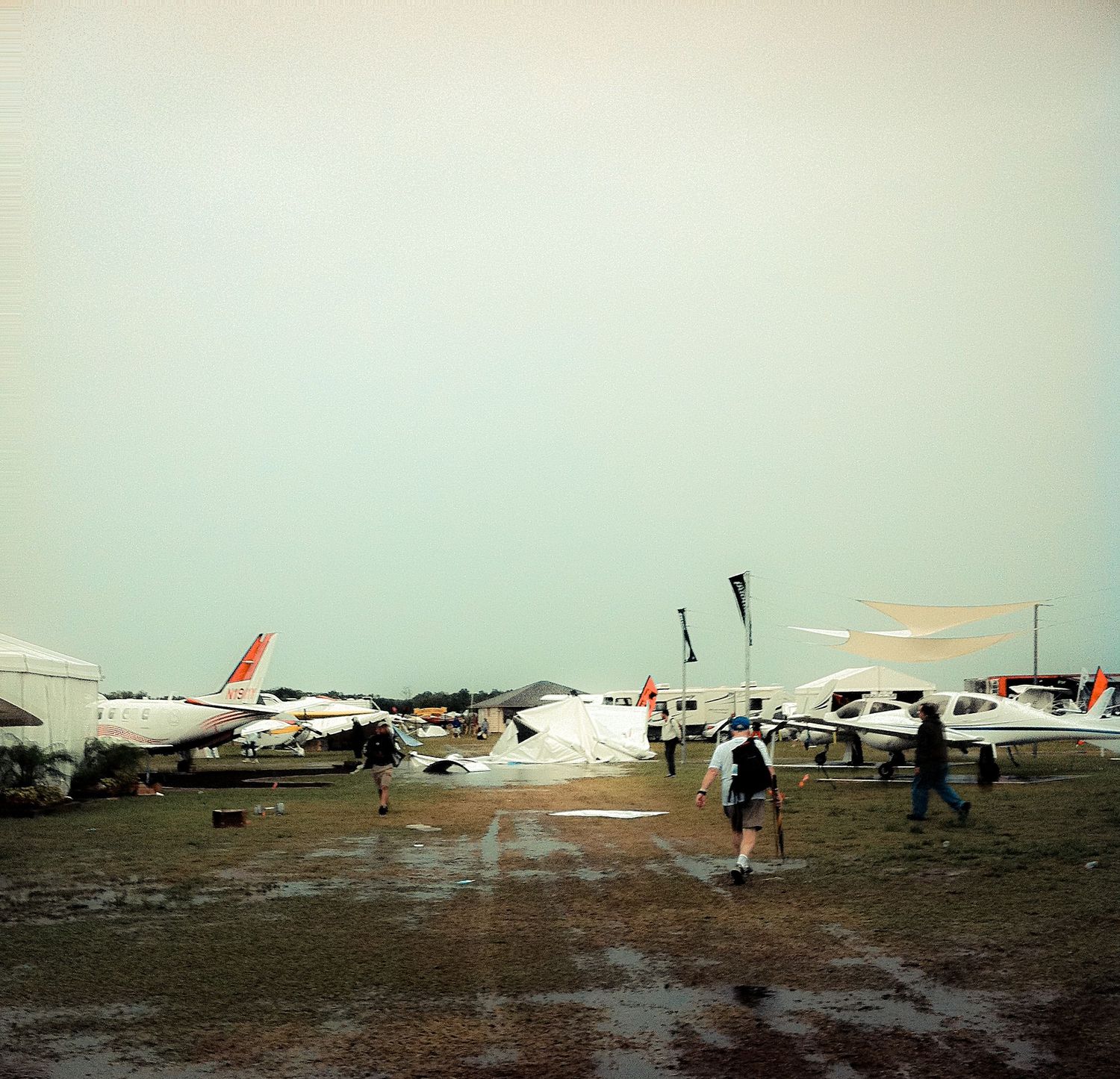 People walk past small aircraft parked on a muddy airfield during an overcast day.
