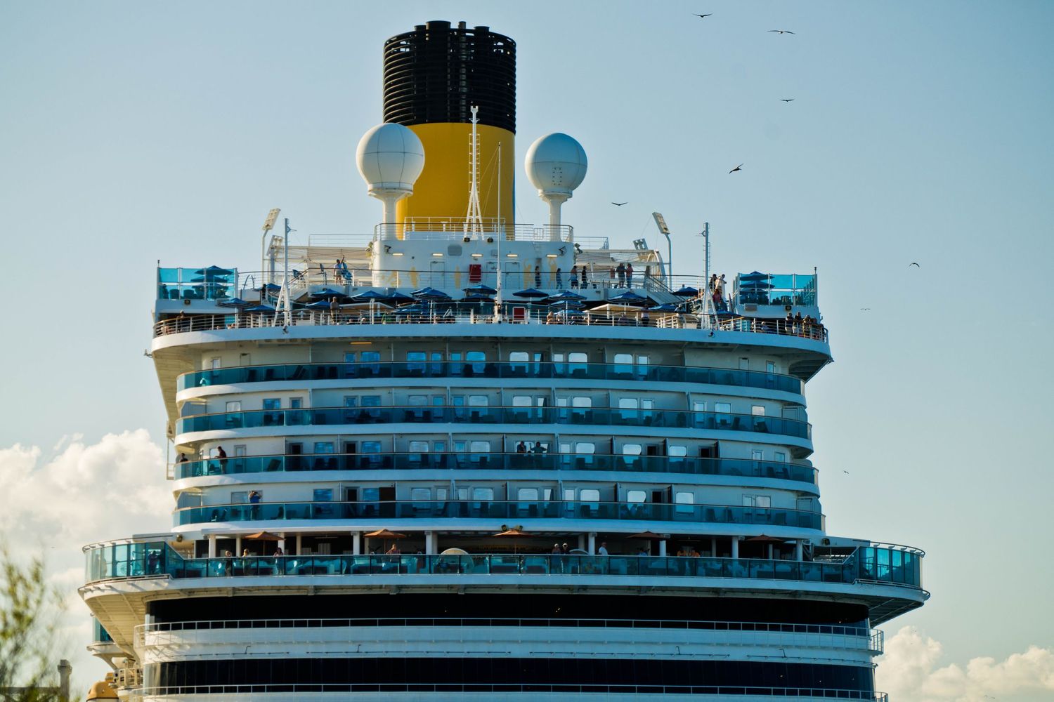Close-up view of a cruise ship's upper decks showing the iconic yellow funnel and white balconies against a blue sky.