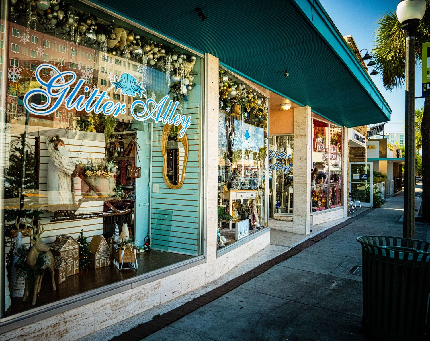 A panoramic view of a shopping street with storefronts and palm trees under blue skies.