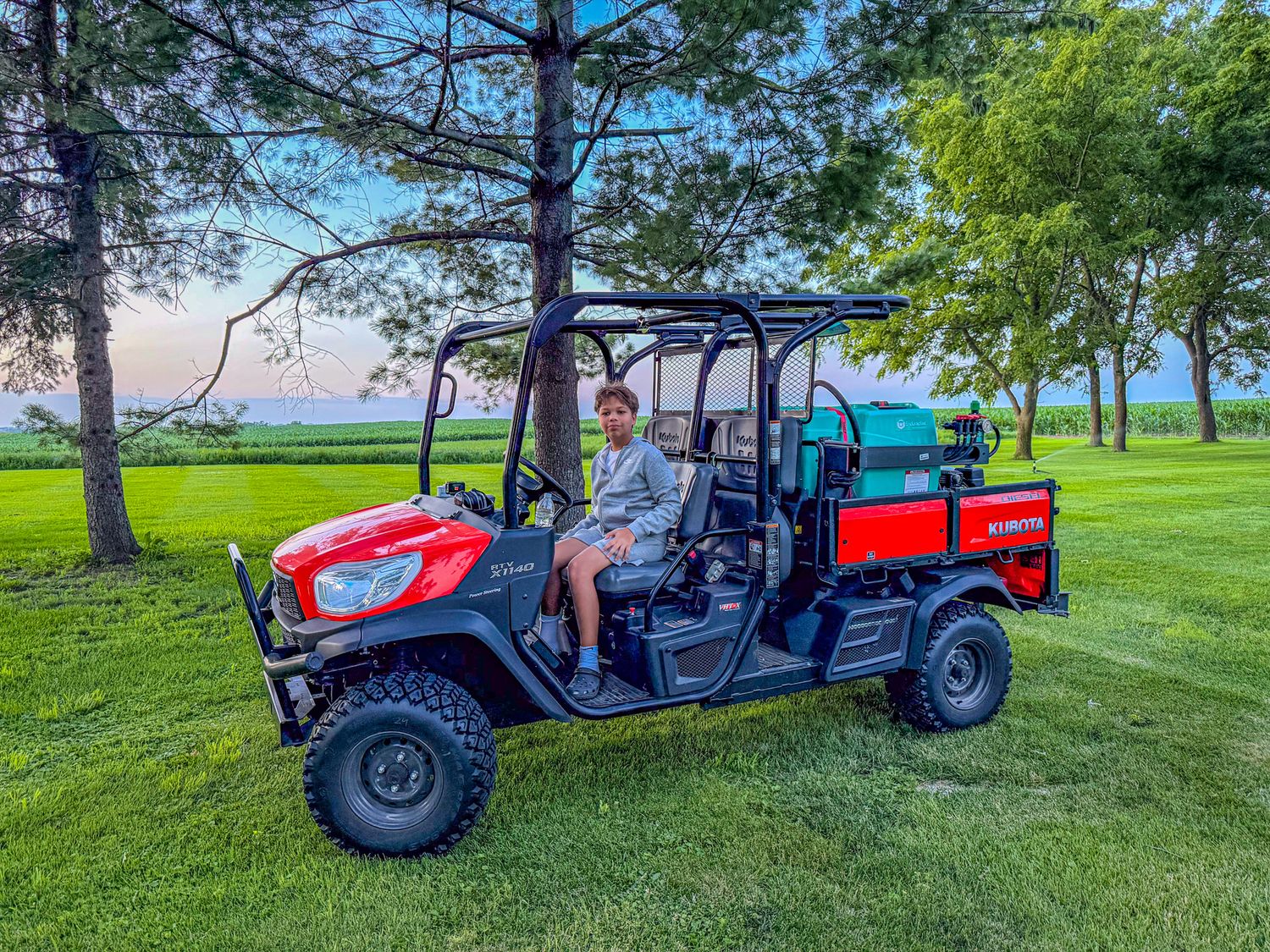 Person driving a red Kubota utility vehicle with a sprayer attachment on a lush green golf course lawn.