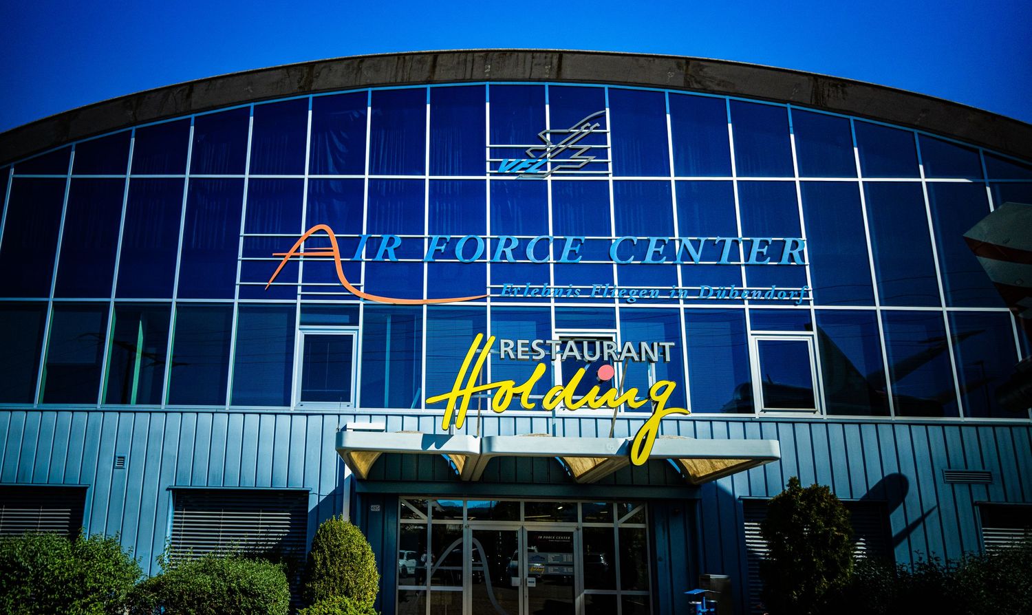 Blue glass-fronted building with neon signage reading Air Force Center and a yellow neon Restaurant Holding sign above the entrance.