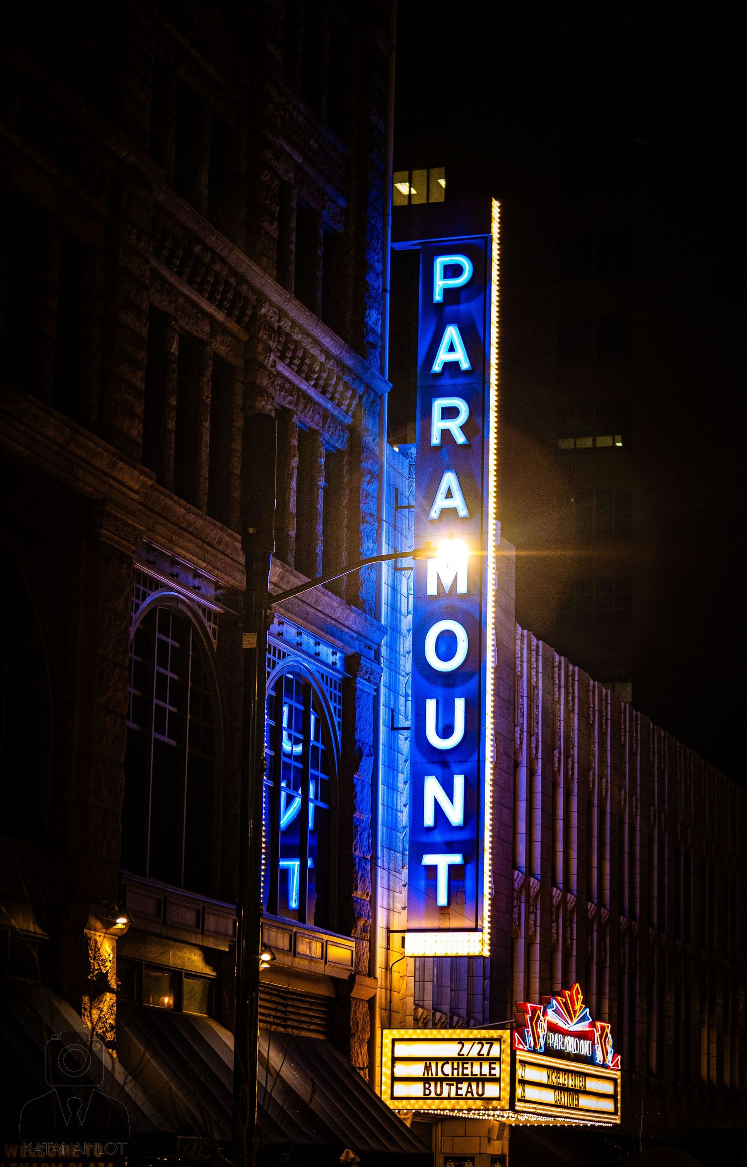 Illuminated neon theater sign letters spelling 'PARAMOUNT' glow blue against dark urban buildings at night.
