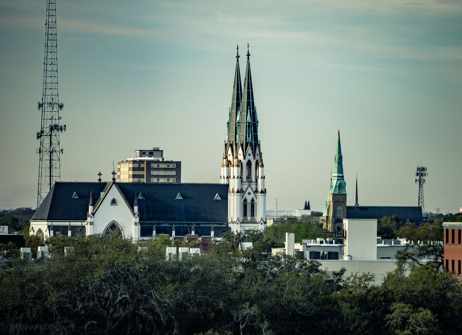 Gothic church spires and towers rise above the Savannah skyline against a blue sky with communication towers visible.