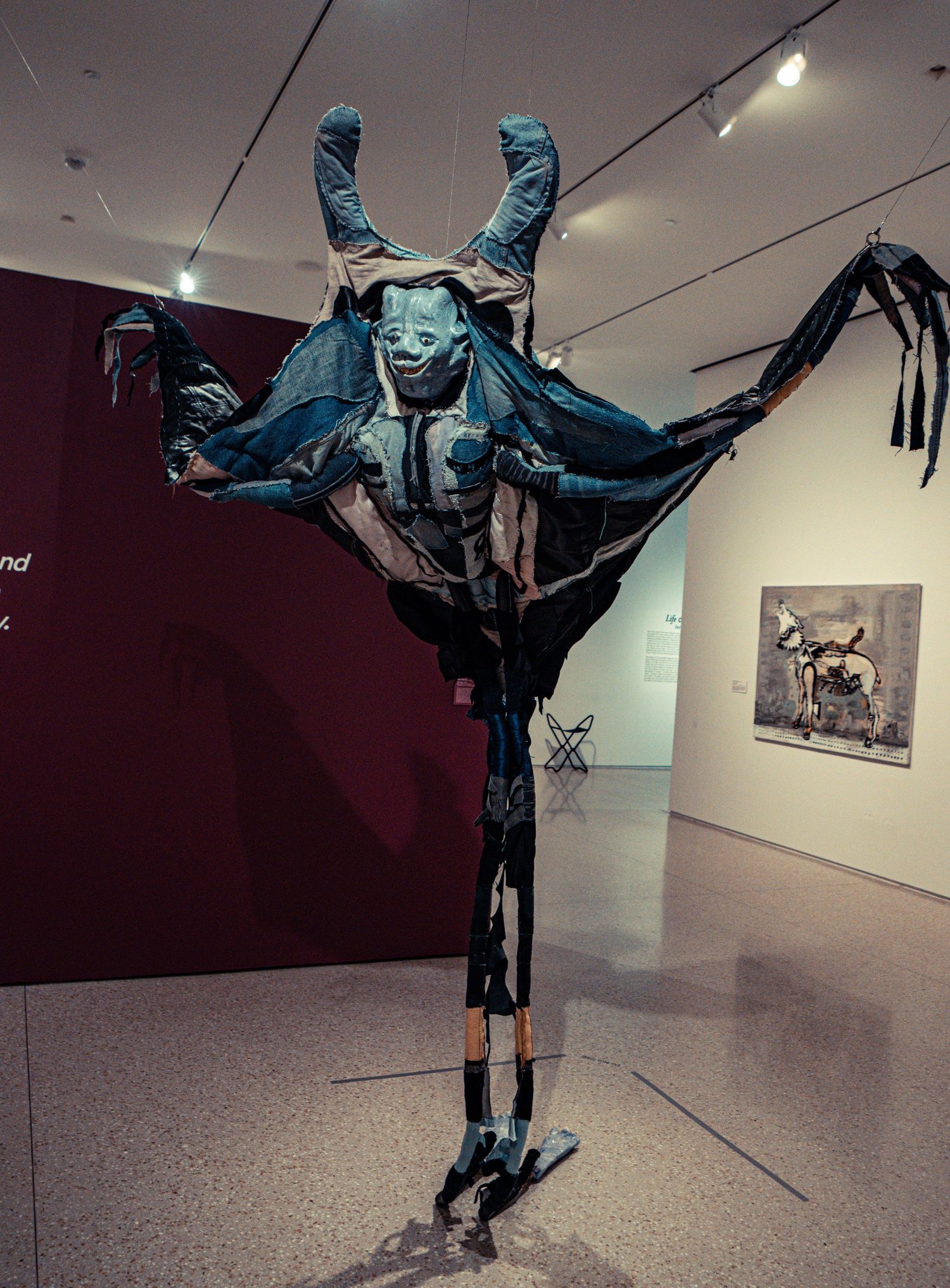 Dramatic sculpture of a horned figure in blue fabric suspended in a gallery space with outstretched limbs.