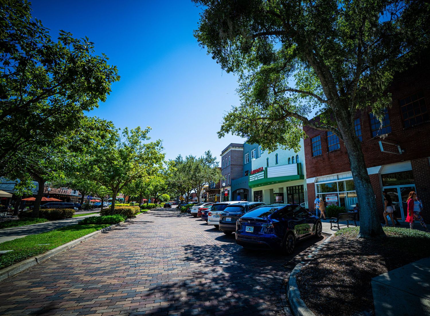 Tree-lined brick walkway with storefronts and parked cars on a sunny day.