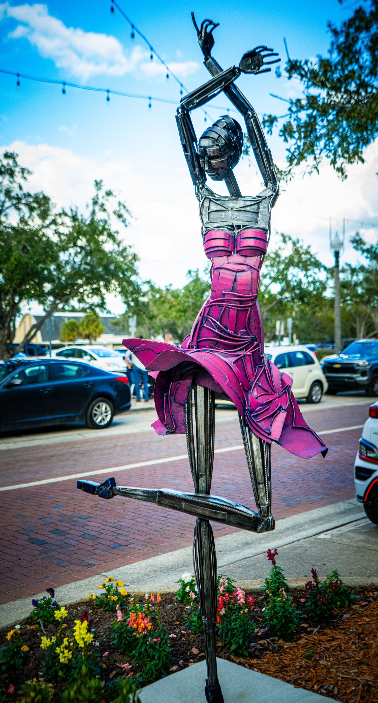 A pink metallic sculpture of a dress form stands in a garden bed with colorful flowers and brick pavers in the background.