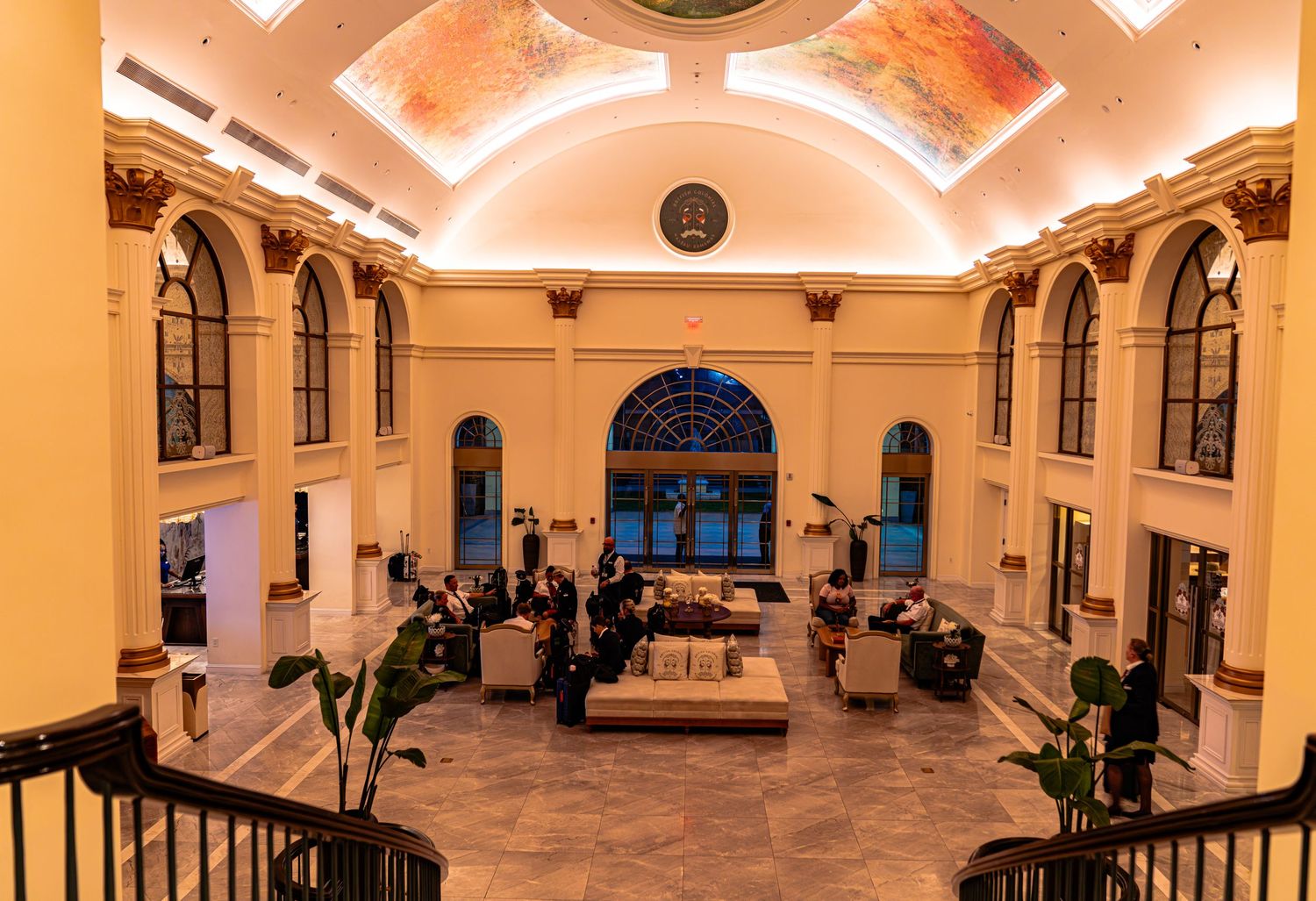 Elegant hotel lobby with arched windows, ornate ceiling mural, warm lighting, and guests relaxing in a grand historic building.
