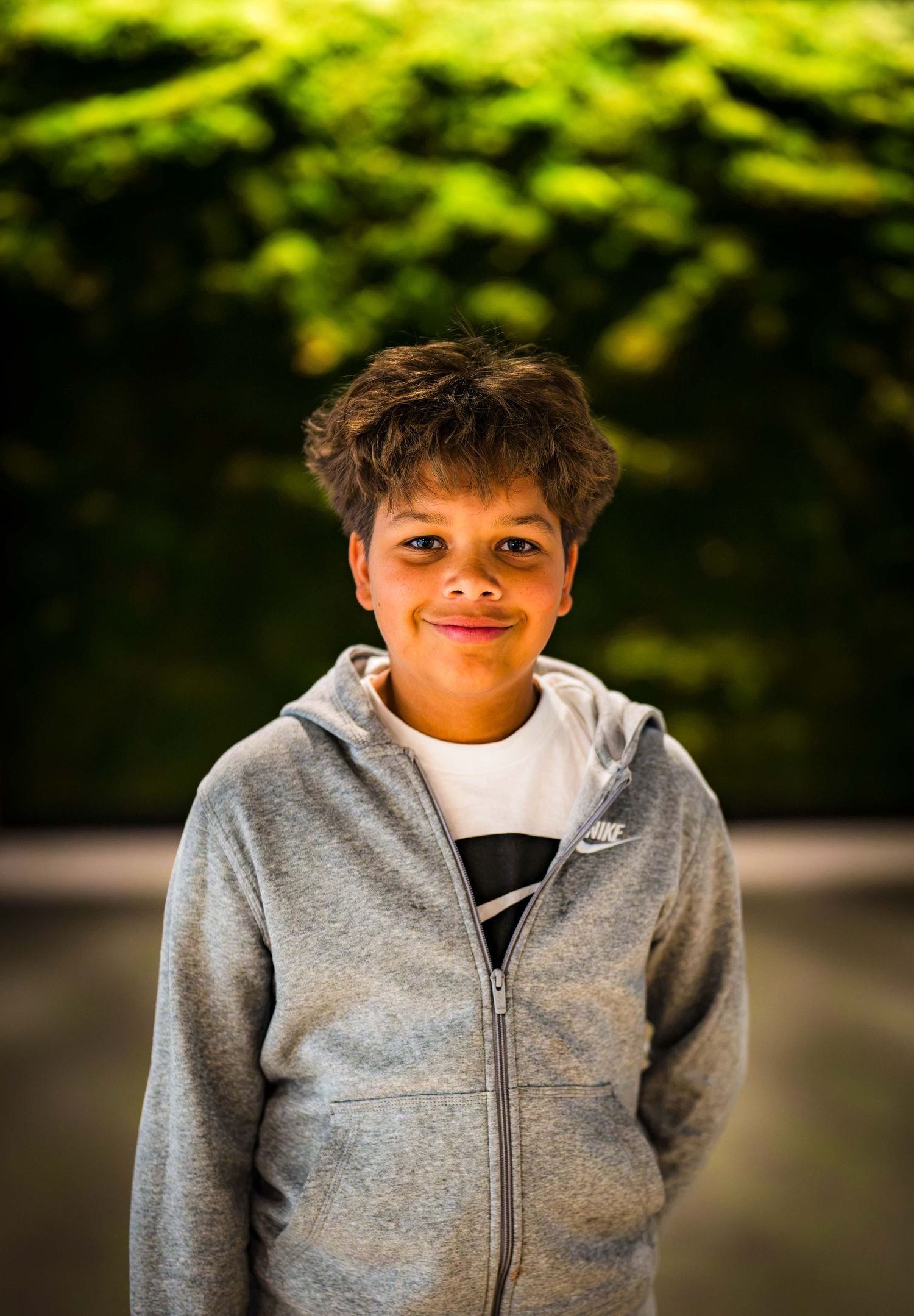 Young boy in gray Nike hoodie smiling outdoors with lush green foliage background.