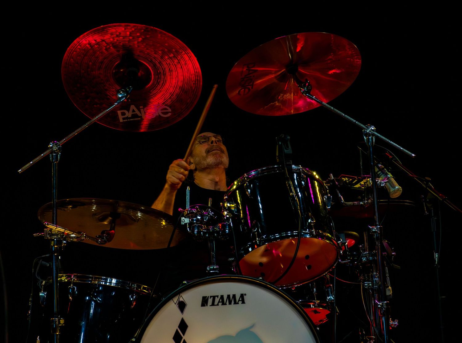 Drummer performing on a Tama drum kit under red stage lighting, mid-play with drumsticks raised.