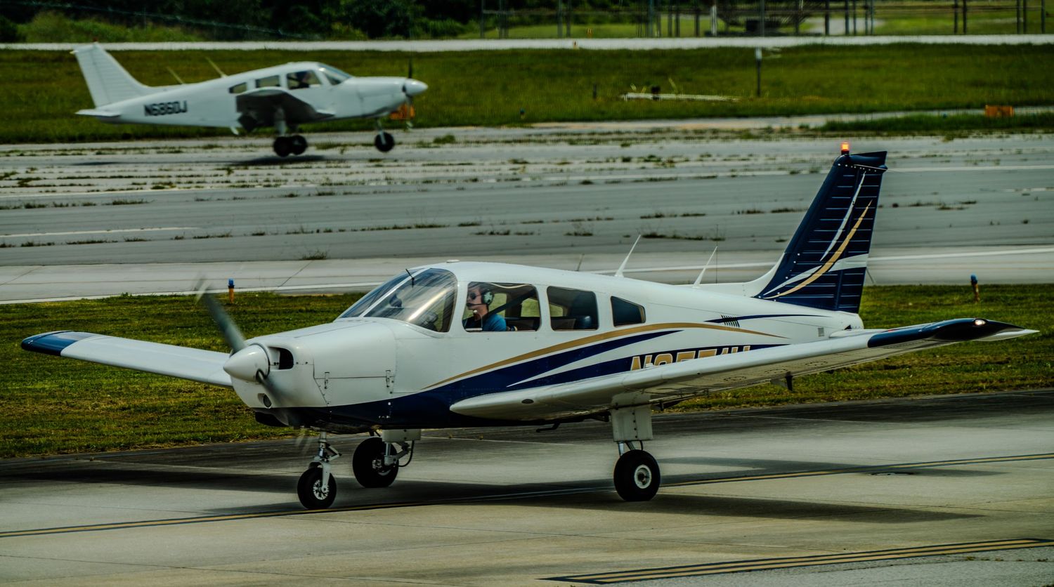 Small white and blue propeller plane taxiing on runway with second aircraft visible in background.