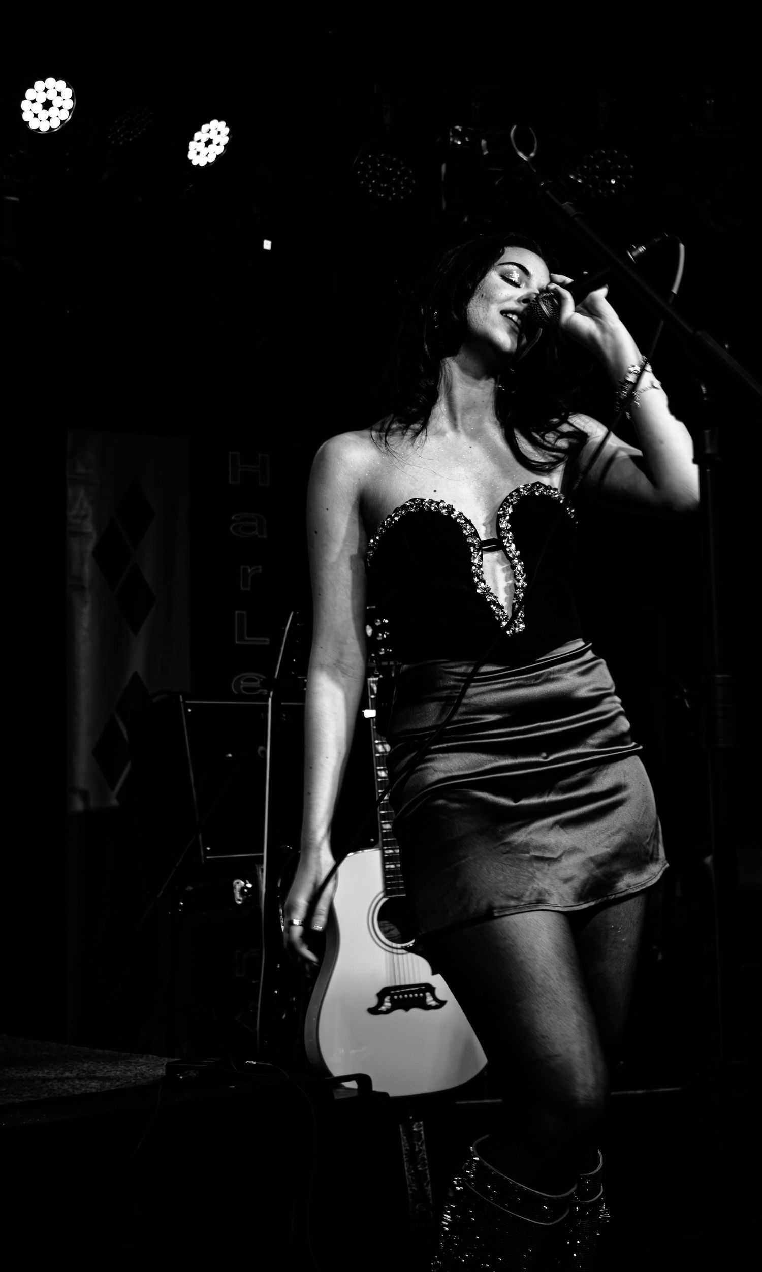 Black and white photo of a woman in a strapless dress holding an acoustic guitar on a dimly lit stage.