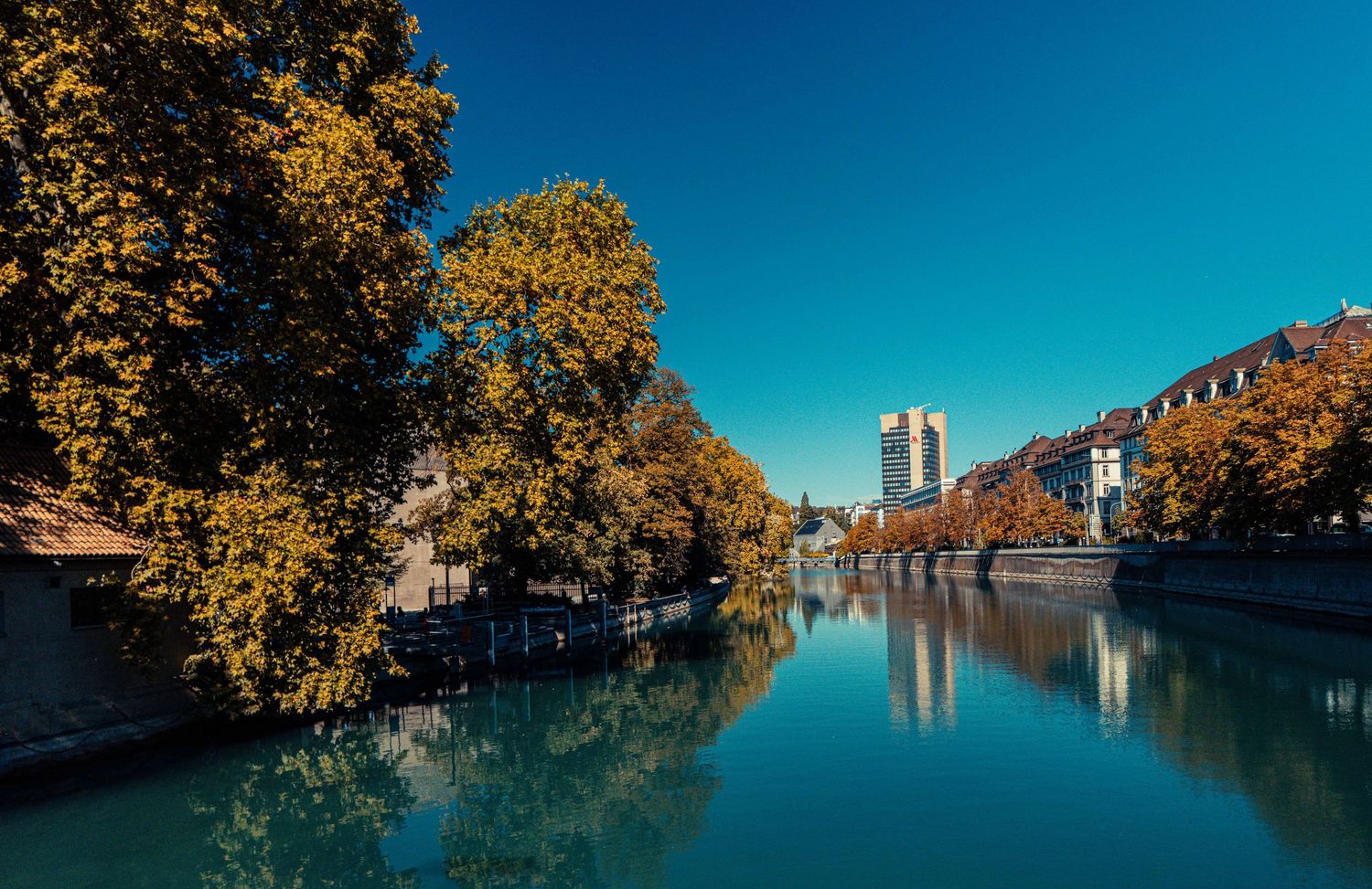 Calm river reflecting golden autumn trees and city buildings under a clear blue sky.