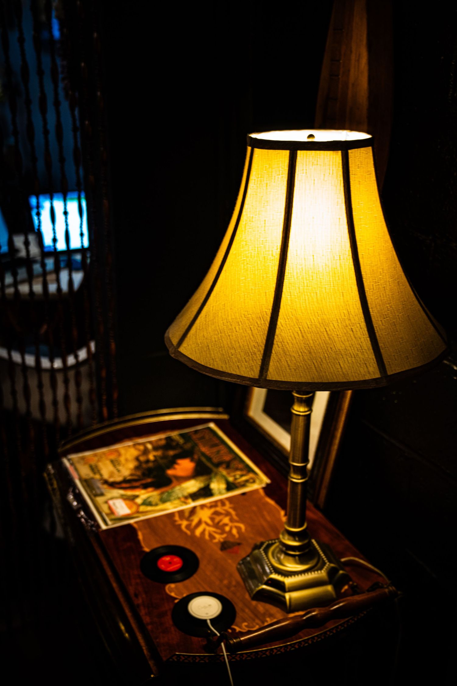 A table lamp with a warm yellow shade illuminates vintage items and records in a dark room.