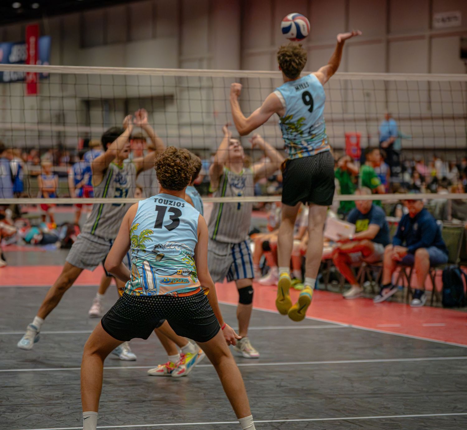 Teen boys playing competitive indoor volleyball, player #9 jumping to spike ball over net during tournament match.