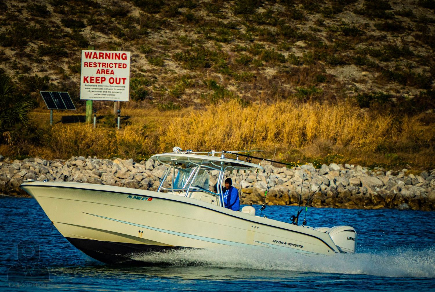 Warning signs posted along a rocky shoreline with boats visible in the distance on a sunny day.