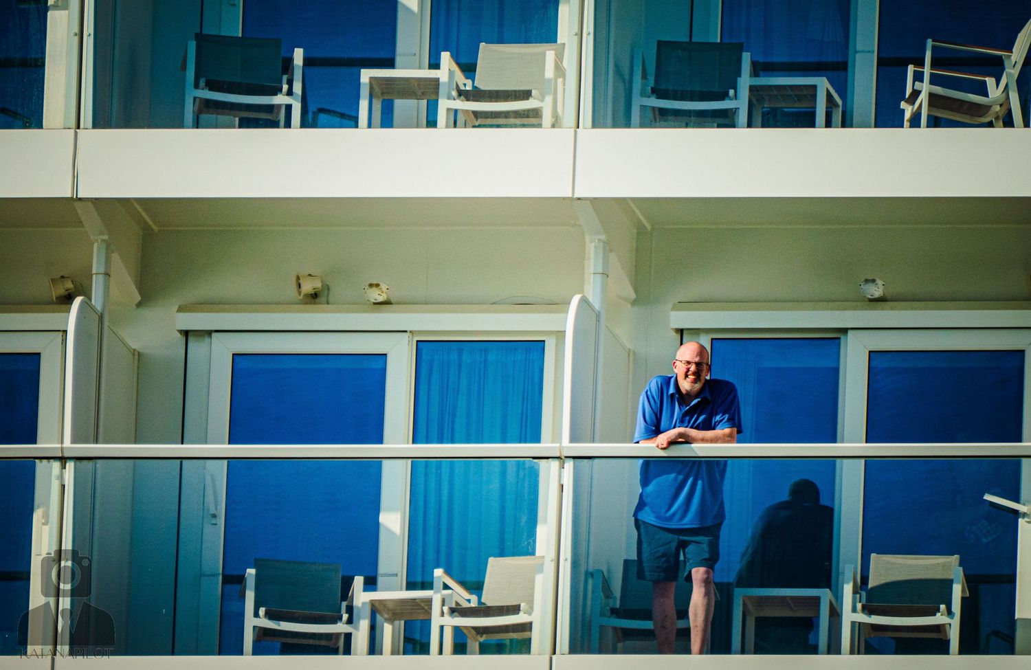 Person standing on a cruise ship balcony against bright blue glass and white architecture.