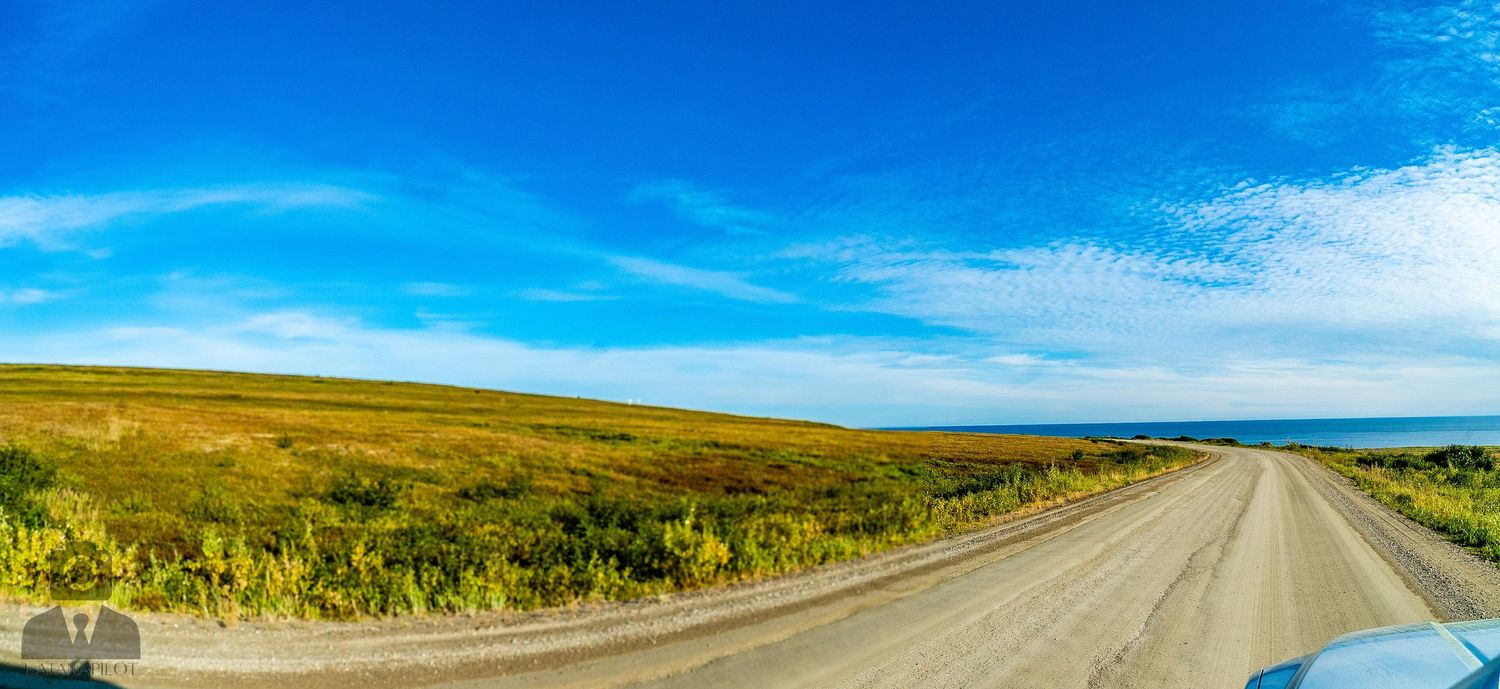 Long dirt road stretching through vast green tundra landscape under blue sky.