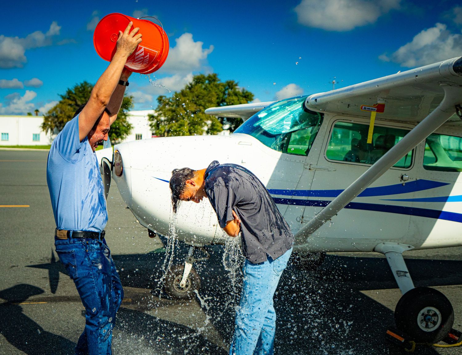 Man pouring red bucket of water on another person near a small white Cessna airplane on a sunny airport tarmac.