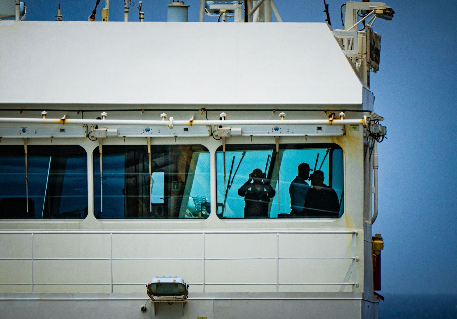 Two crew members visible through bridge windows of a large white commercial ship against a blue sky.