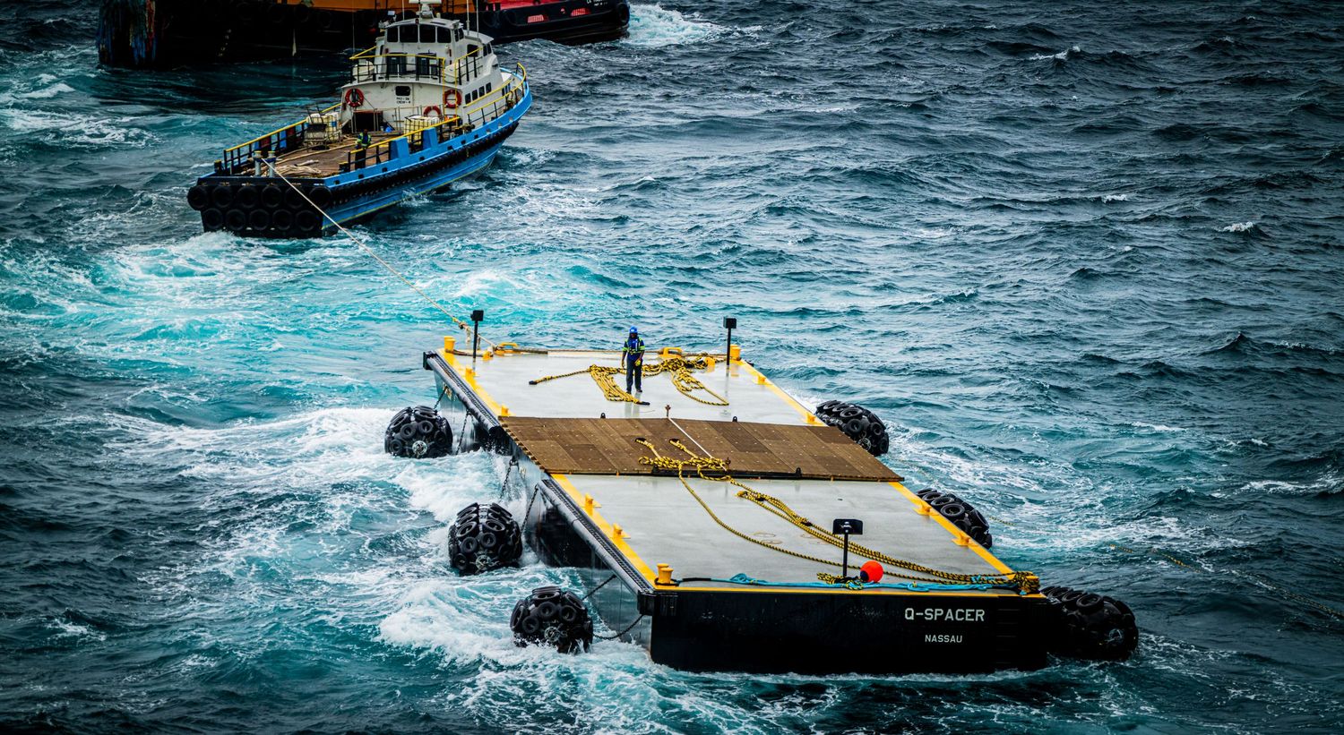 Yellow and black offshore barge navigating rough ocean waves with a blue tugboat nearby.