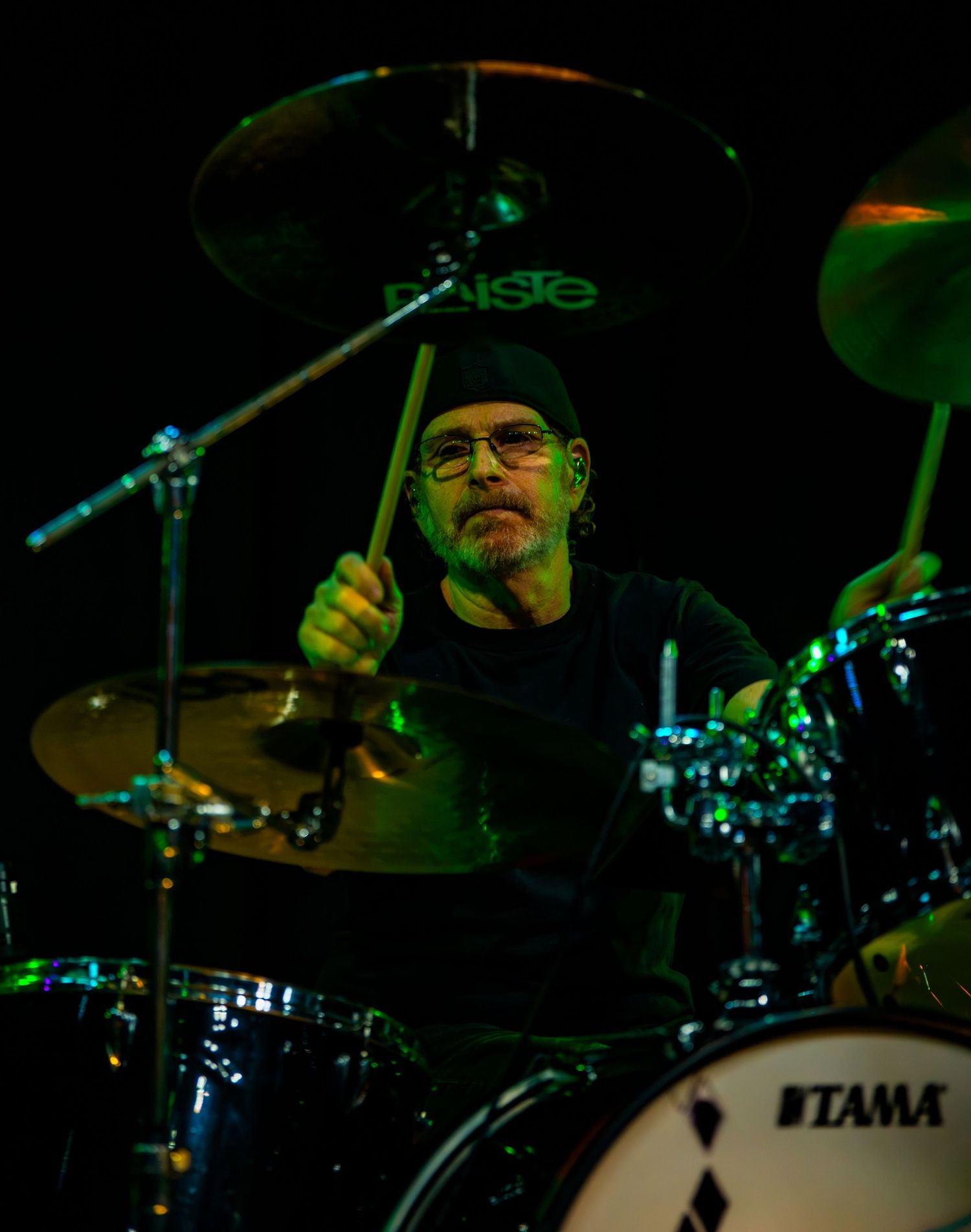 A bearded drummer plays Tama drums with Paiste cymbals on a dark stage, lit by green and blue lights.