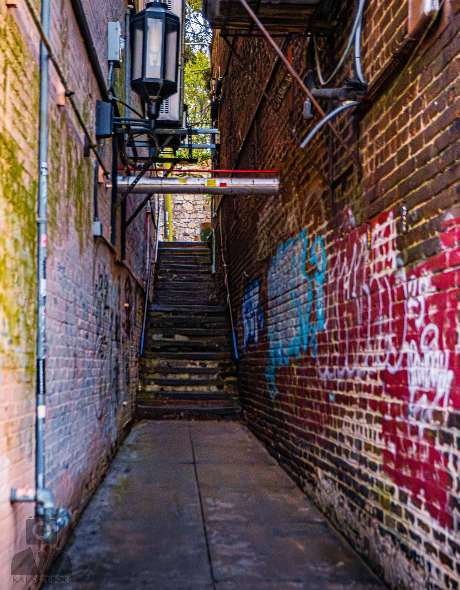 Narrow brick alleyway with graffiti walls leads up steep stairs while a vintage lantern hangs overhead in an urban setting.